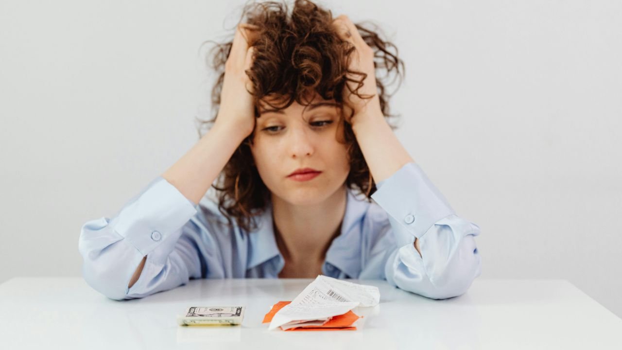Young woman holding her head in stress while reviewing bills and counting money