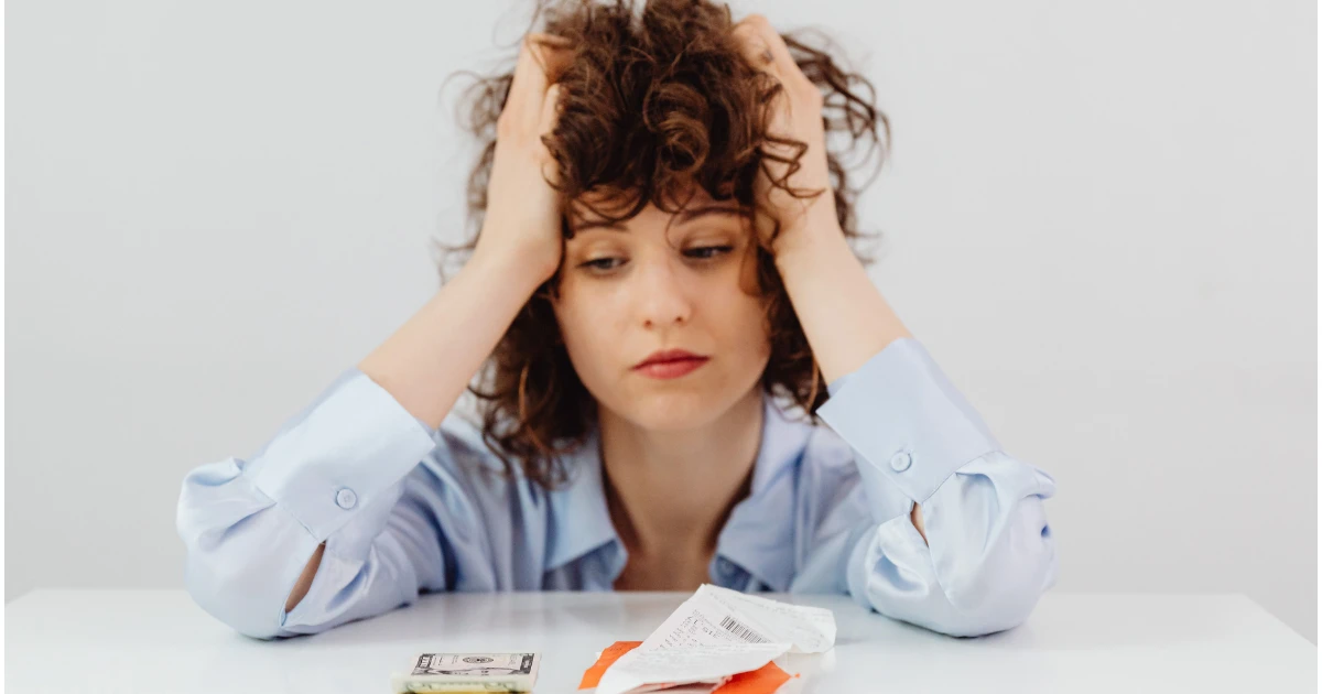 Young woman holding her head in stress while reviewing bills and counting money